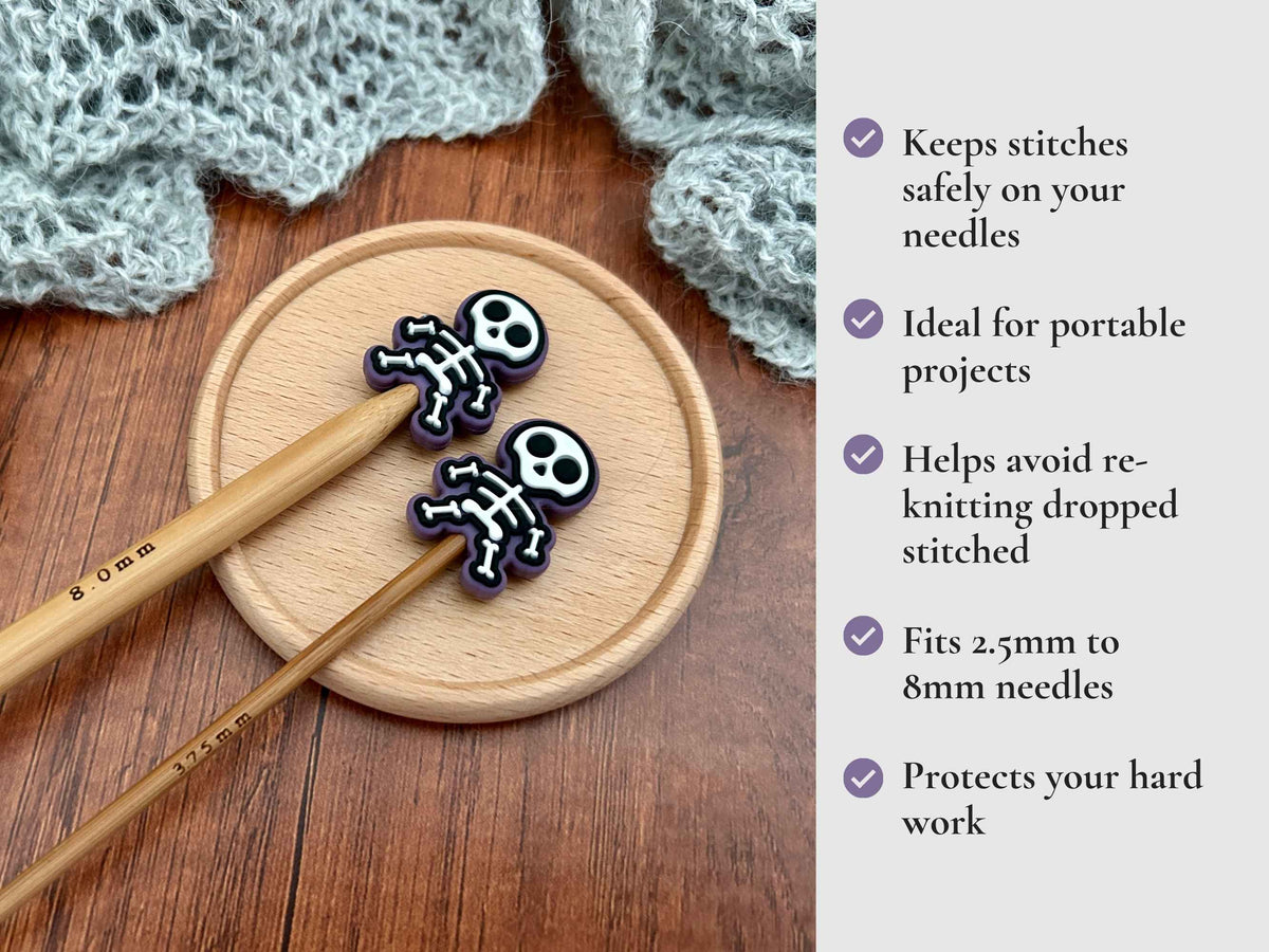 Two small skull-shaped needle guards with black and white bone designs are shown resting on wooden knitting needles on a round wooden tray, with a knitted gray fabric in the background and a text list of benefits on the right. Keeps stitches on needles. Ideal for portable projects. Helps avoid re-knitting dropped stitches. Fits mm to 2.5mm to 8mm needles 
