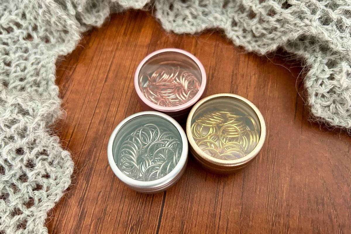 Three small round metal containers with clear lids, each filled with 40 snag free knitting stitch markers. The tins and corresponding stitch markers are steel coloured, rose gold-coloured, and gold-coloured. They are,arranged on a wooden surface with a gray knitted textile in the background.