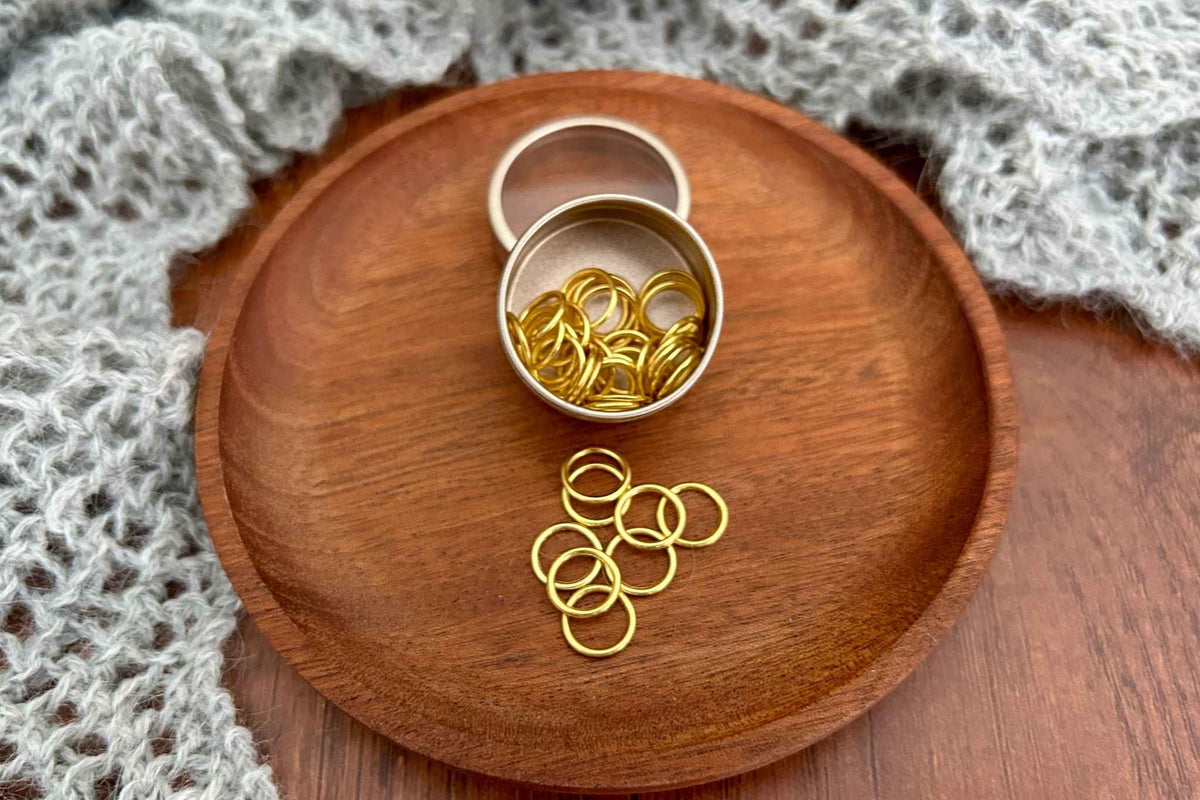 Gold metal ring snag free stitch markers  displayed in an open round tin and scattered on a wooden tray, with a gray knitted textile in the background.