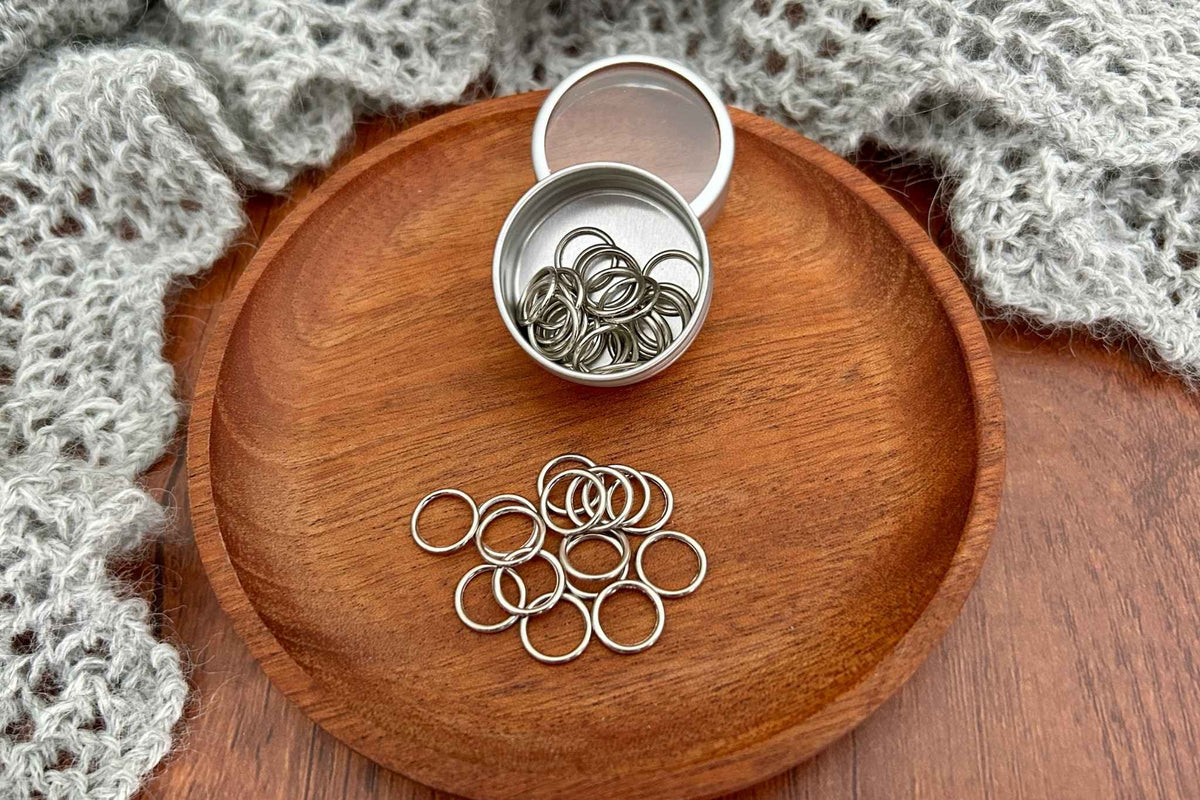Silver metal snag free stitch markers scattered on a round wooden tray, with an open small metal tin containing more ring dings above them. The tray is styled with a gray knitted textile on a wooden surface.