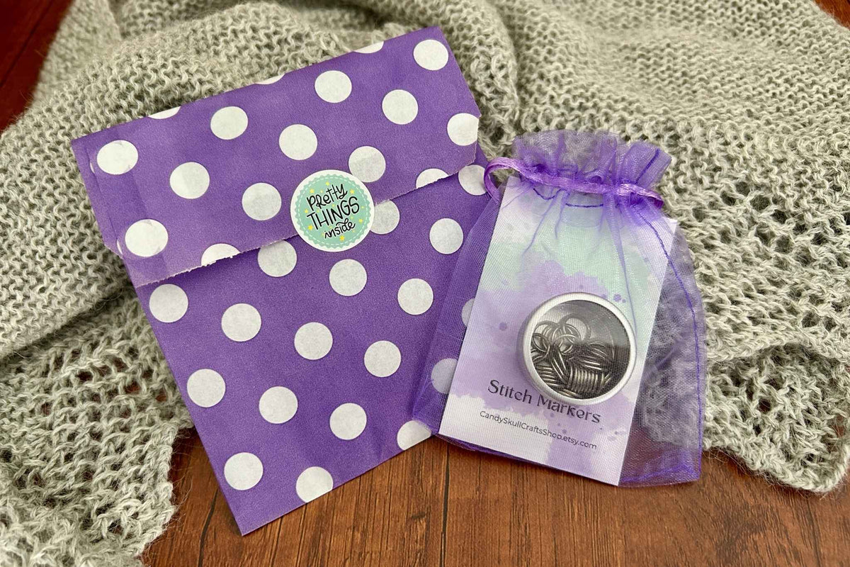 A purple and white polka dot envelope with a 'Pretty Things Inside' sticker, next to a small purple organza gift bag containing a steel coloured set of knitting stitch markers set on a grey knitted blanket over a wooden surface.