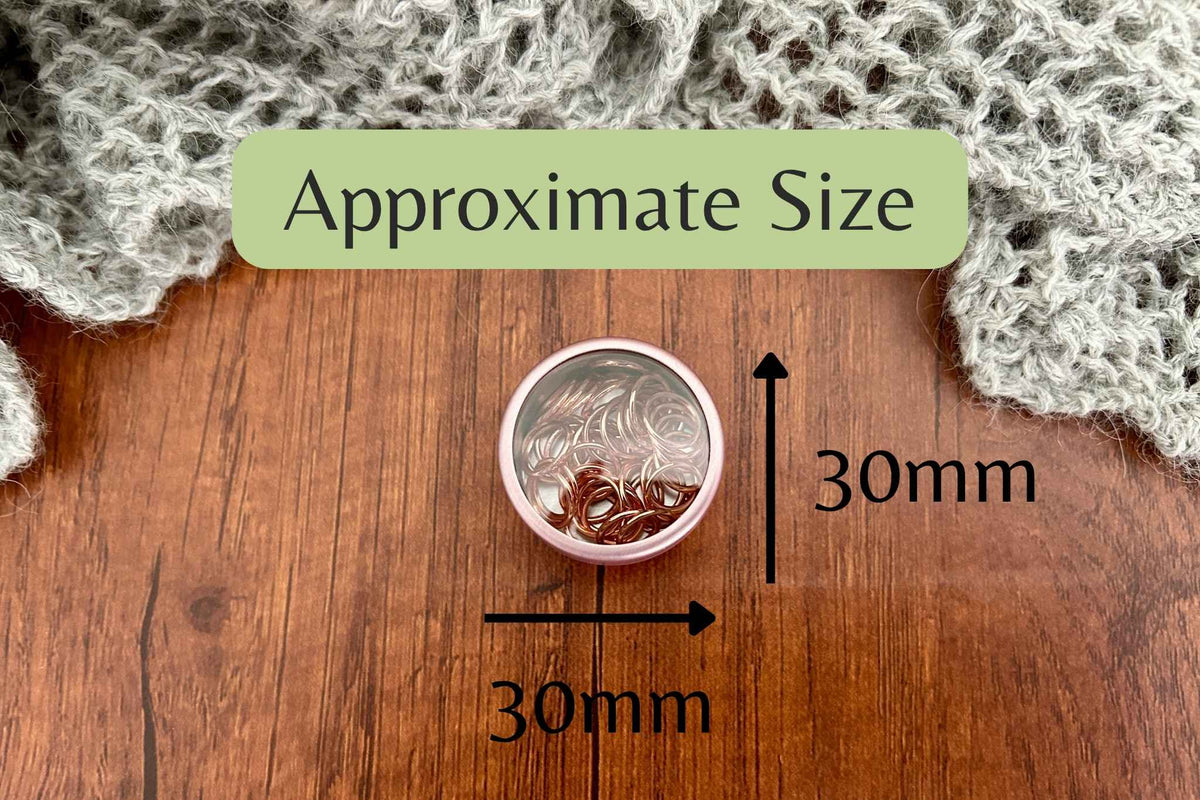 A small round metal stitch marker pot with a clear lid, shown on a wooden surface with a green label reading 'Approximate Size' and arrows marking dimensions as 30mm by 30mm and 30mm tall. Knitted gray fabric is visible at the top corners.