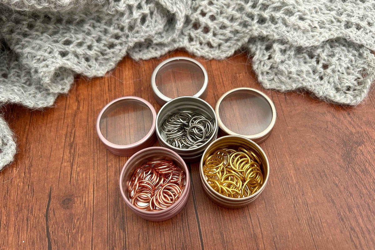 3 small round metal tins filled with knitting stitch markers in steel, rose gold and gold. The tins are arranged on a wooden surface with a gray knitted blanket in the background.