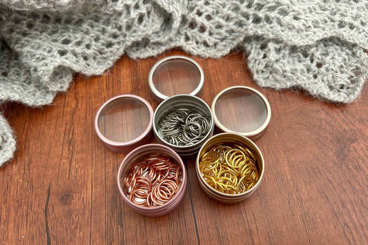 3 small round metal tins filled with knitting stitch markers in steel, rose gold and gold. The tins are arranged on a wooden surface with a gray knitted blanket in the background.