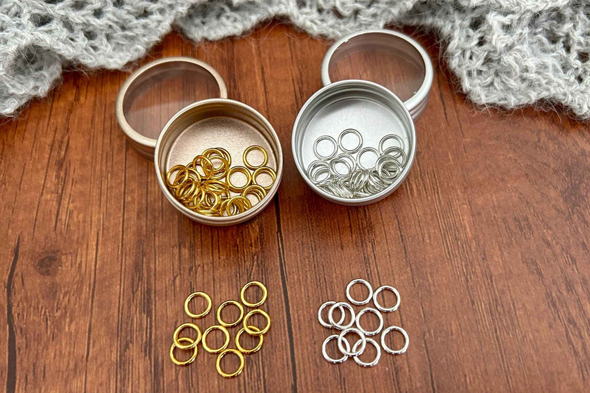 Two small round metal pots filled with snag free knitting stitch markers ring dings, one gold-toned and one silver-toned, arranged on a wooden surface with loose matching rings in front and a gray knitted fabric at the top.