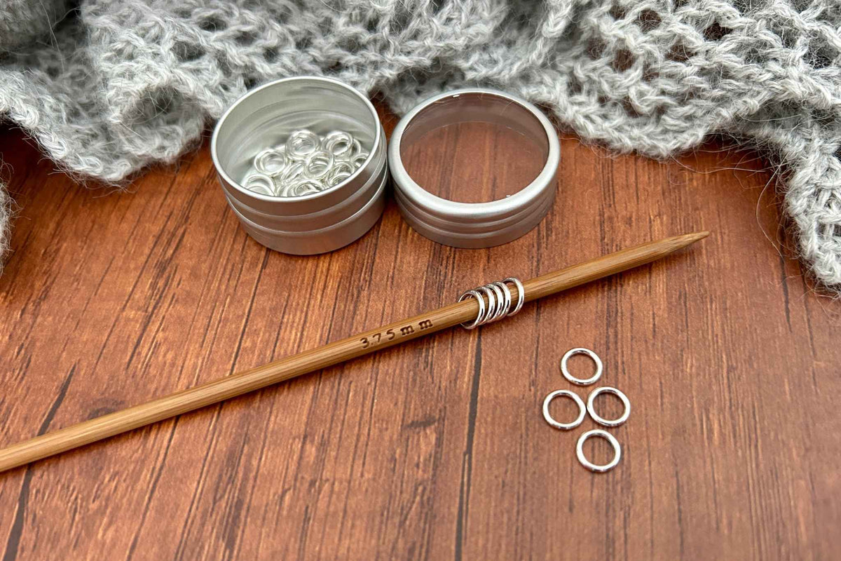 A wooden 3.75mm knitting needle with silver metal snag free stitch markers, displayed on a wooden table next to two small metal containers of silver jump rings and loose rings, with a gray knitted blanket in the background.