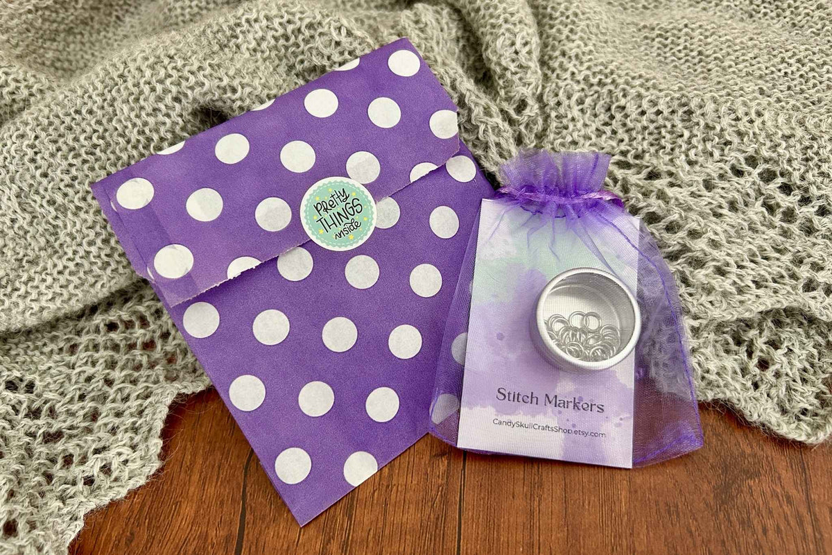 A small silver metal stitch marker tin in a clear purple tulle pouch sits beside a purple polka-dot envelope on a wooden surface with a knitted blanket behind it. The package label reads 'Stitch Markers' and shows the brand candyskullcrafts