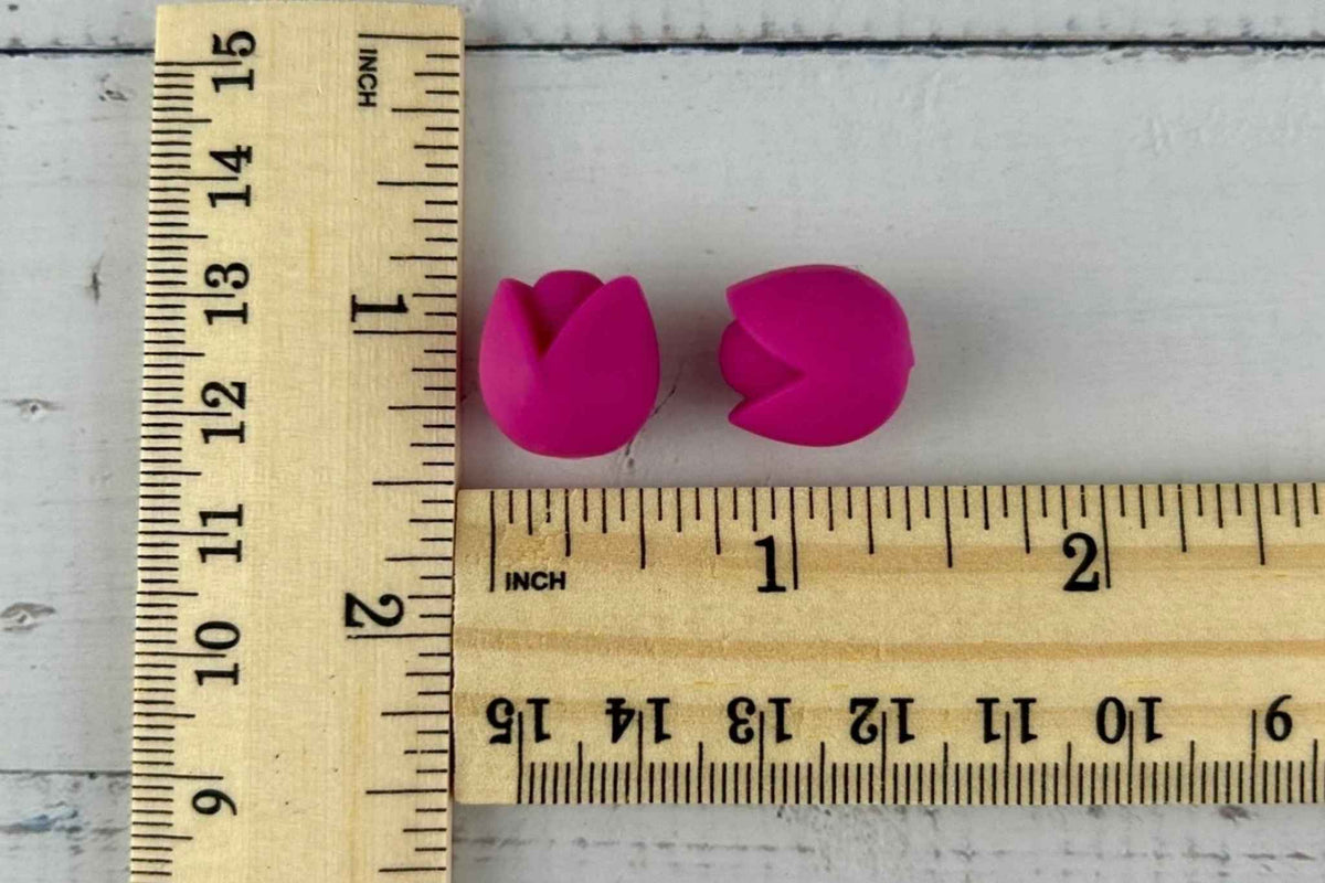 Two small rose pink tulip-shaped knitting needle stoppers shown on a white surface with wooden rulers for scale. Approx size 15mm by 15mm