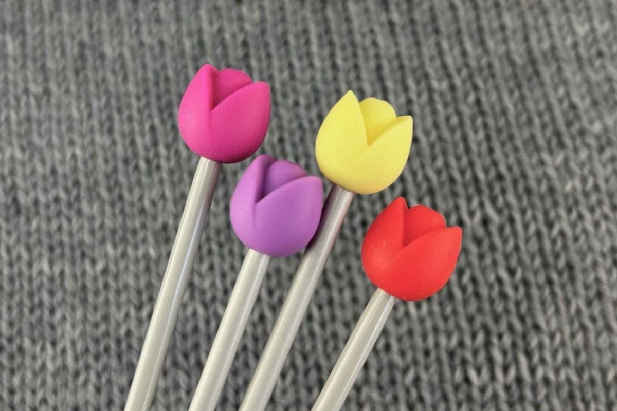 Four tulip-shaped knitting needle stoppers in pink, purple, yellow, and red, attached to metal knitting needles, displayed on a gray textured background.