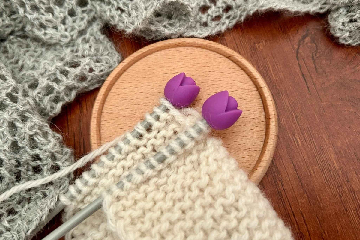 Two purple tulip-shaped knitting needle stoppers resting on a round wooden coaster, with a cream-coloured knitting. The stoppers are preventing the knitting from sliding off.  A gray knitted textile is visible in the background.
