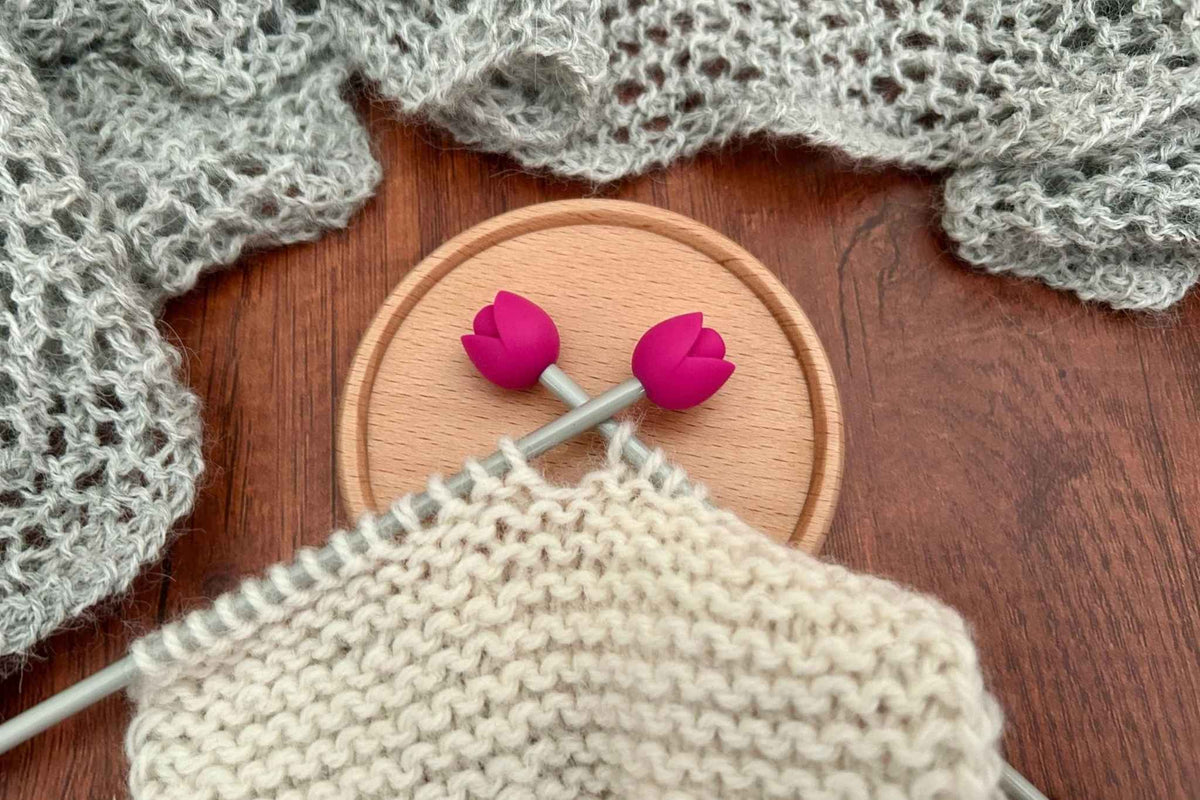 2 rose pink tulip-shaped stitch stoppers preventing cream knitting from sliding off the metal needles. A gray knitted scarf or shawl is draped around the scene.