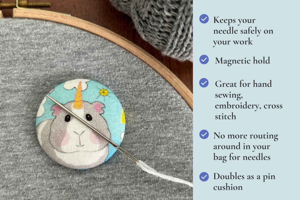 A round fabric magnetic needle minder or pin cushion featuring a cute cartoon unicorn guinea pig design, shown on a gray fabric surface next to a wooden embroidery hoop and a needle. A light blue text panel on the right lists product features with checkmarks. keeps your needle safely on your work. Magnetic hold great for hand sewing, embroidery, cross stitch. No more routing around in your bag looking for needles. Doubles as a pin cushion.
