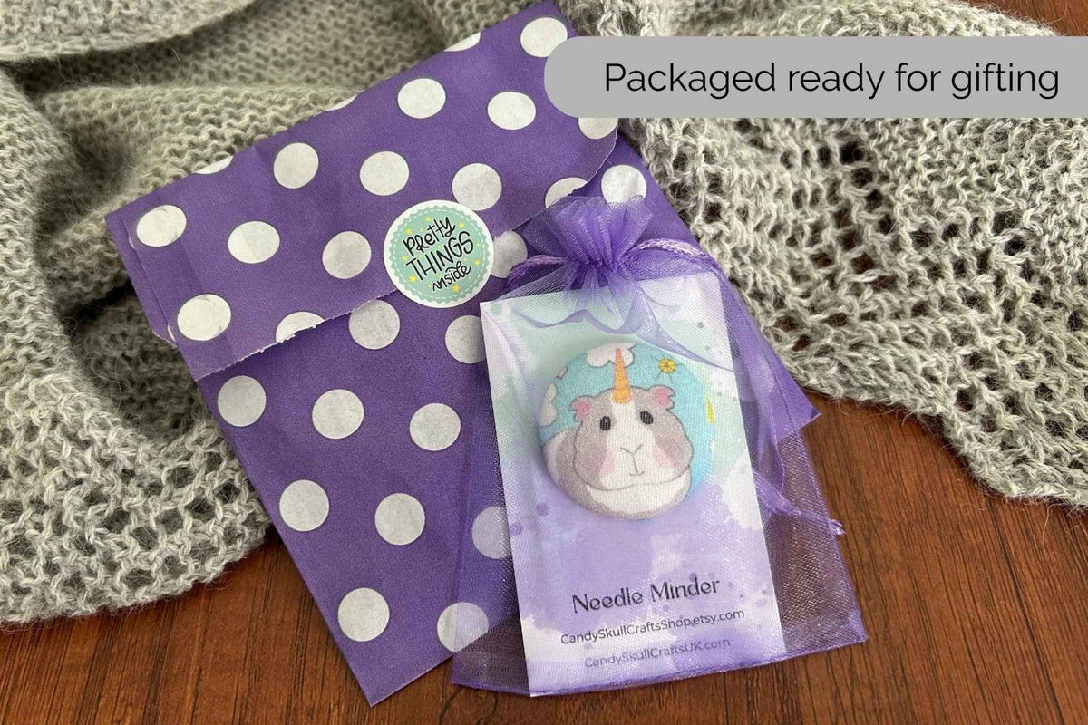 A packaged magnetic fabric needle minder featuring a unicorn guinea pig in an organza mesh bag. Nest to it is the packaging of a purple and white polka dot bag. 