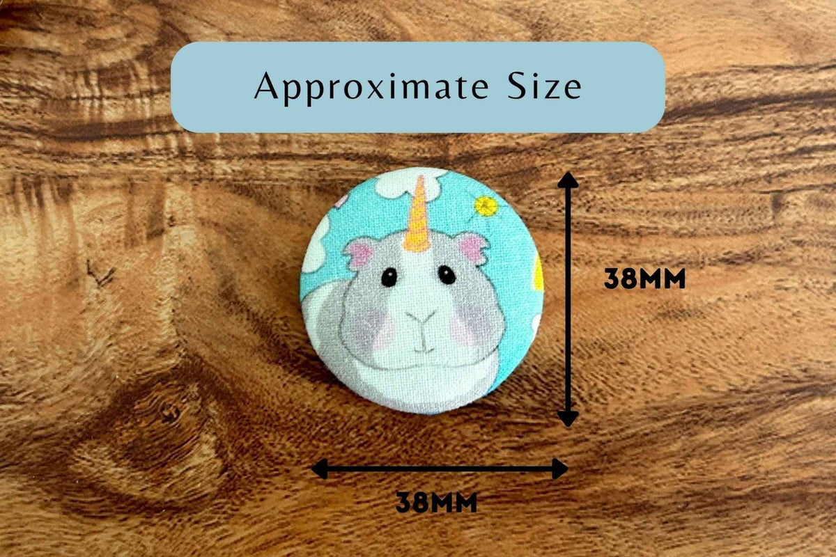 A round magnetic needle minder eaturing a cute cartoon unicorn guinea pig on a light blue background, shown on a wooden surface with measurement arrows and labels indicating approximately 38mm by 38mm.