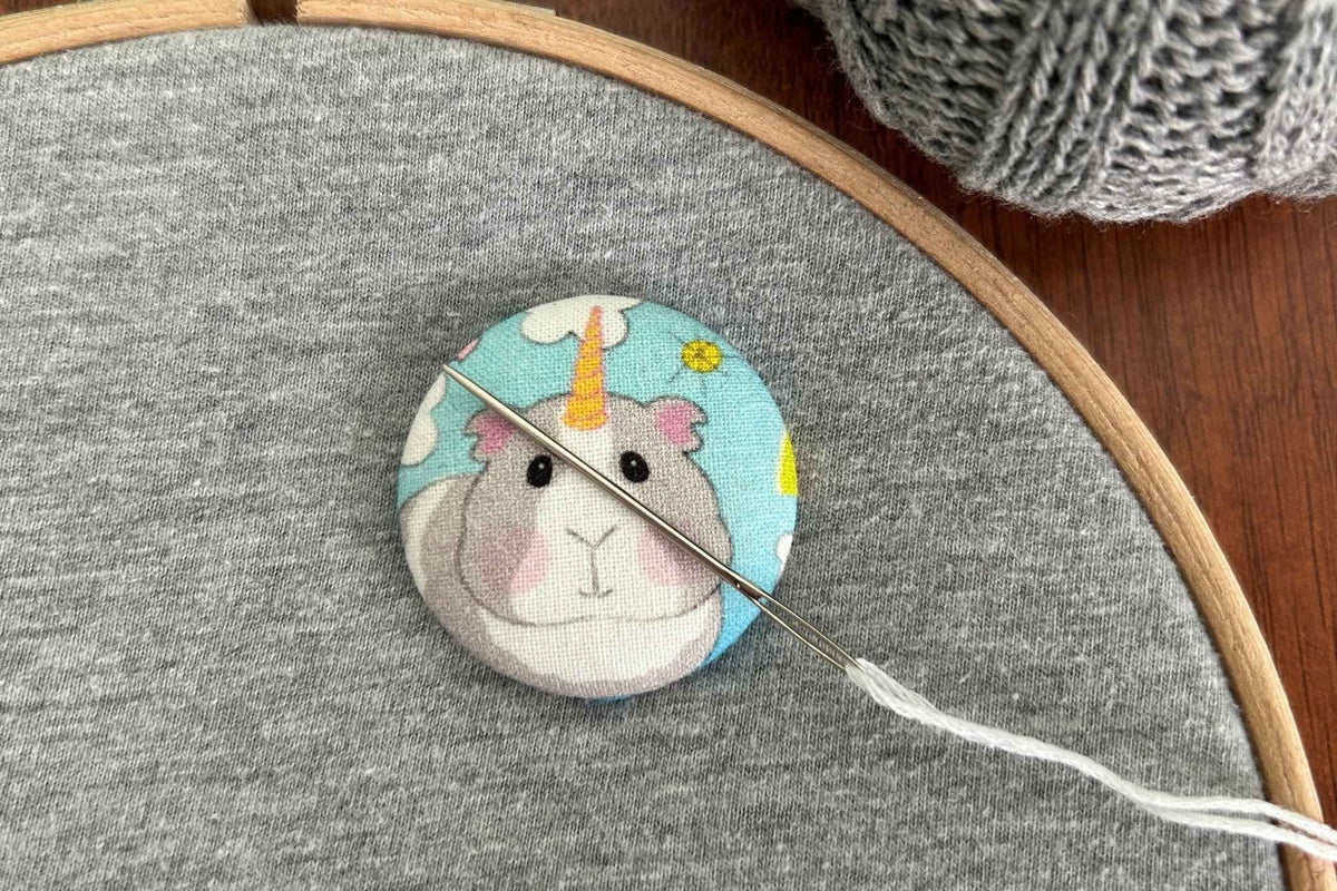 A close-up of a circular fabric needle minder featuring a cute cartoon unicorn guinea pig on a light blue background, placed on gray fabric inside a wooden embroidery hoop with a needle and thread.