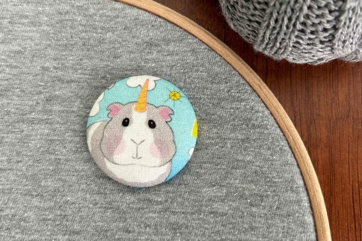 A round fabric magnetic needle minder featuring a cute cartoon unicorn guinea pig unicorn design, displayed on a gray fabric hoop with a wooden frame and a ball of gray yarn on a wooden table.