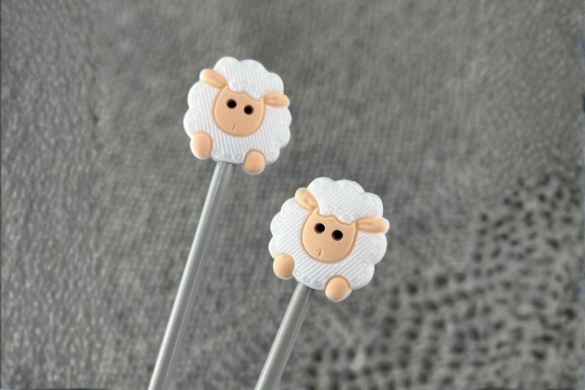 two white sheep shaped needle stitch savers on metal needles displayed on a gray knitted textile

