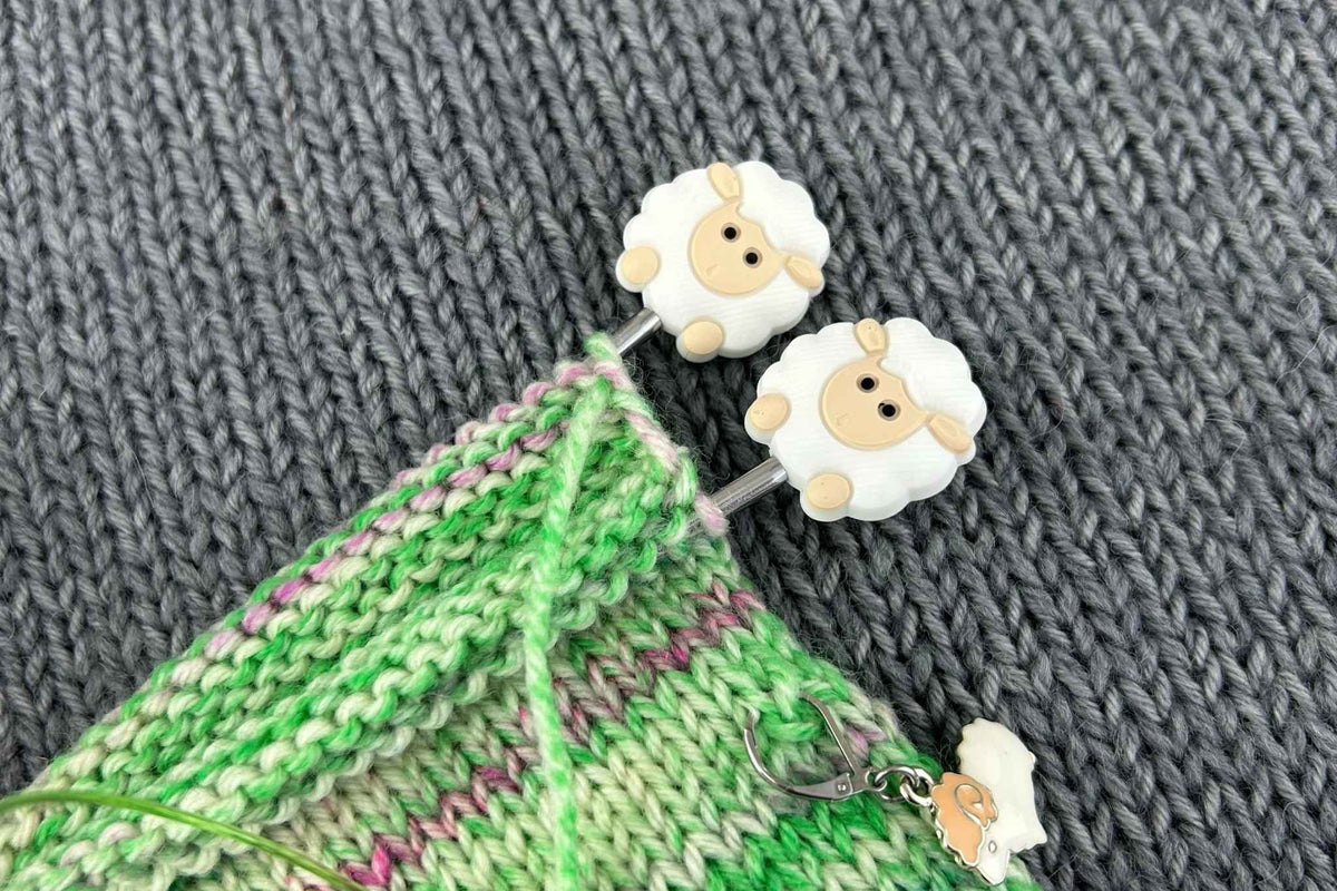 Two white sheep-shaped knitting stitch savers with beige faces are shown on a green and pink knitted swatch, set against a dark gray knitted background.