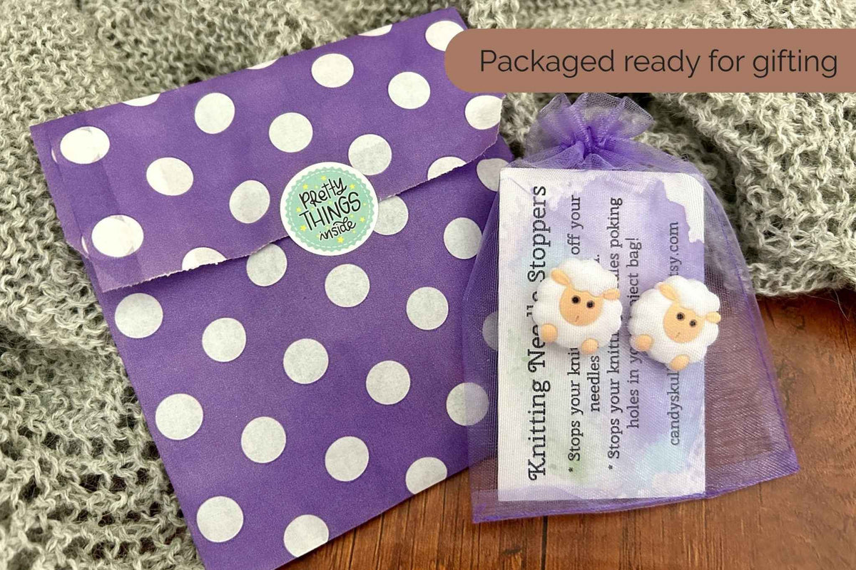 A purple polka dot envelope and a small clear mesh gift bag containing white sheep shaped needle stoppers, with an instruction card, styled on a wooden surface with a gray knit blanket. 
Text reads "packaged ready for gifting"